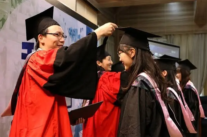 Un profesor con túnica académica roja y negra ajusta la borla del birrete de una estudiante graduada durante una ceremonia oficial.